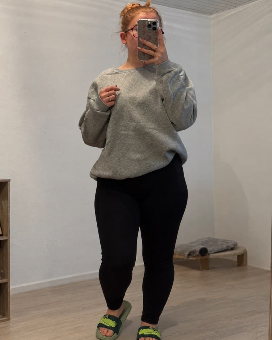 Une cliente portant les claquettes devant un grand miroir, avec un sweat gris et un legging noir.